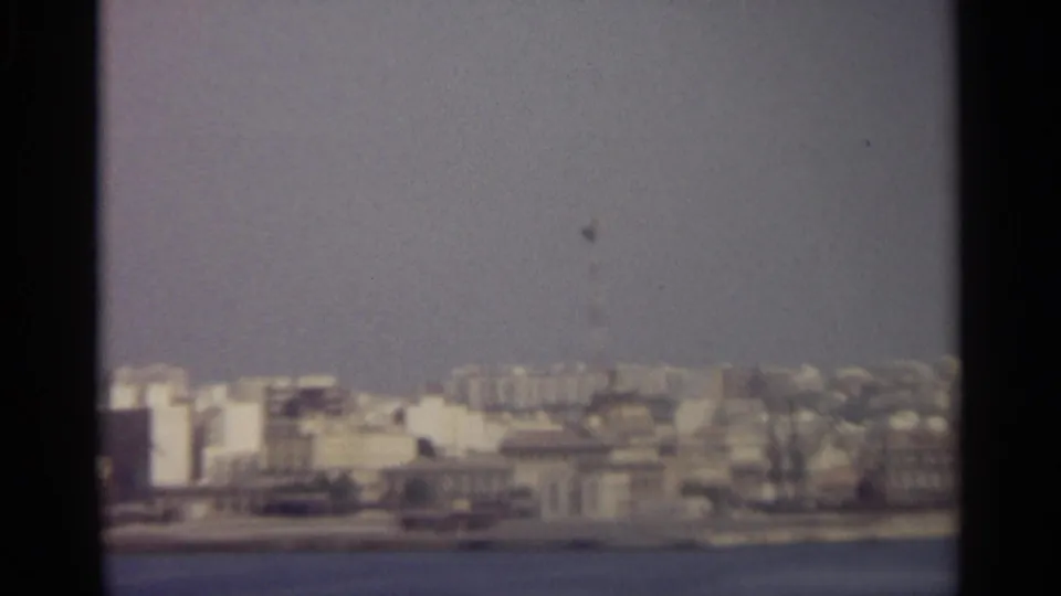 Soft 8mm view of a dense Sicilian waterfront city under a pale blue sky, seen from across the water.
