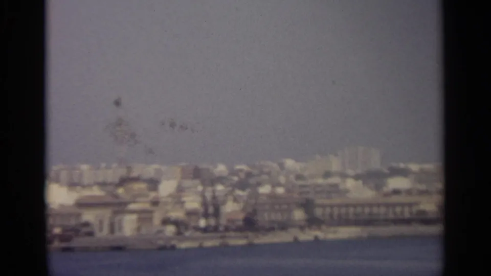 A hazy Sicilian waterfront skyline with clustered buildings and a tall tower rising near the right side.