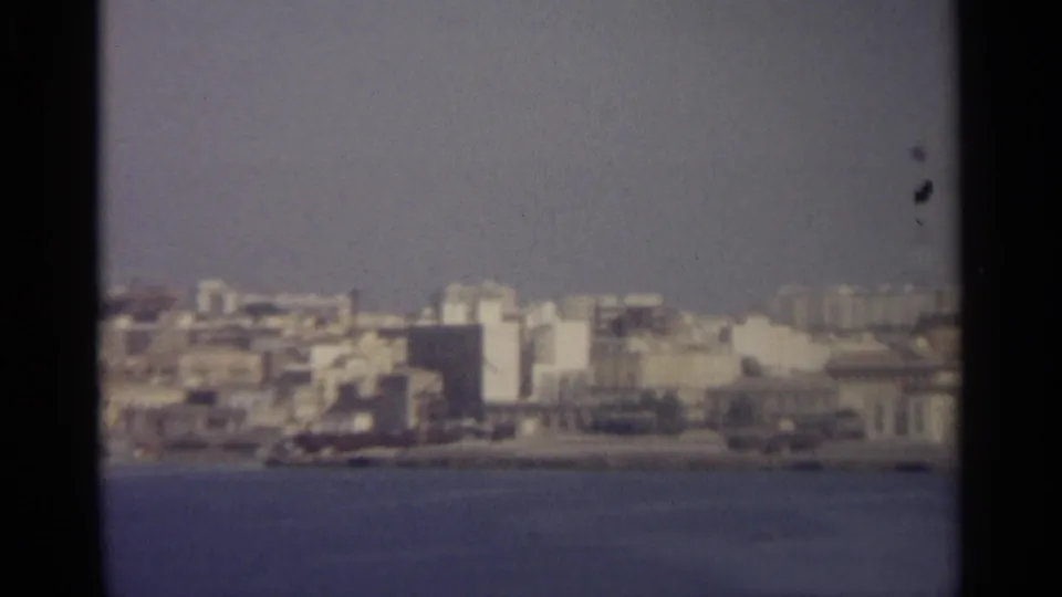 Grainy 8mm frame showing pale buildings packed along a waterfront, with open water in the foreground.