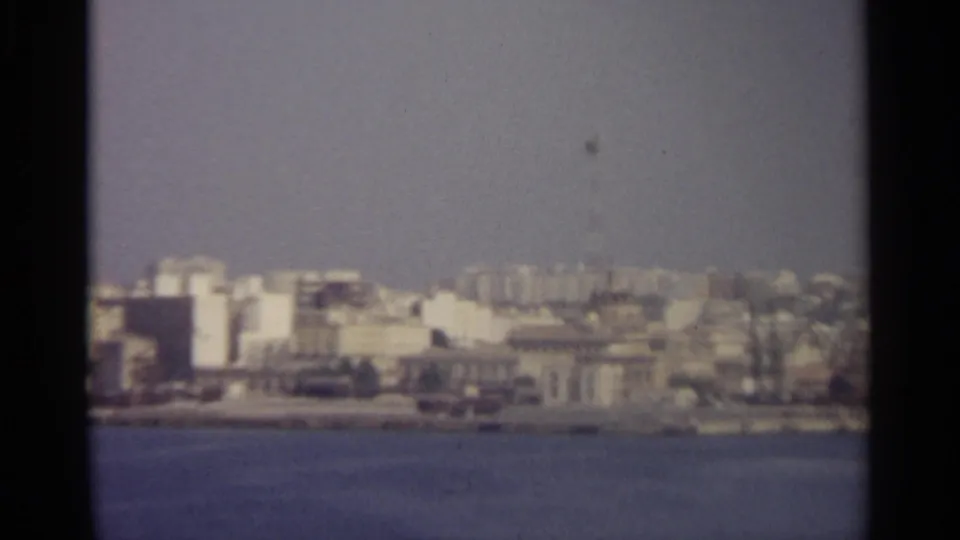 Distant Sicilian shoreline with clustered urban buildings, soft focus, and muted blue water in the foreground.