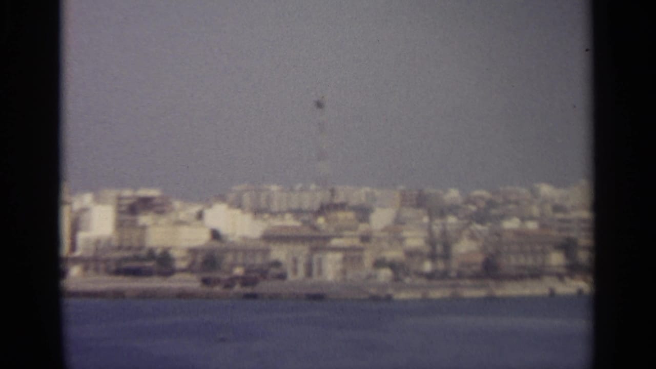 Hazy waterfront city skyline across calm water with dark film edges.