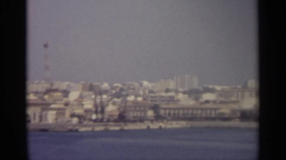 Hazy 8mm view of a Sicilian coastal skyline, with light buildings, a communications tower, and water in the foreground.