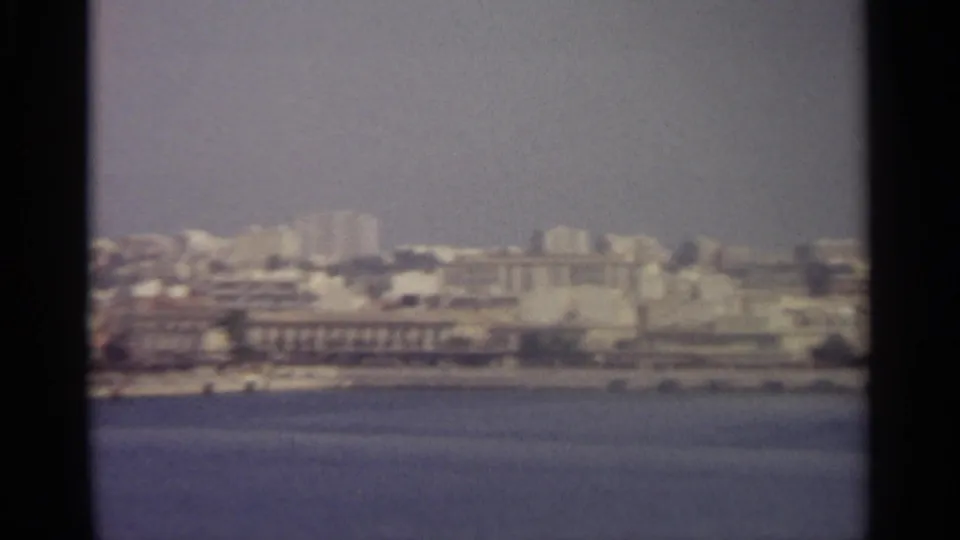 1965 home-movie frame showing a soft, hazy Sicilian waterfront cityscape from across the water.