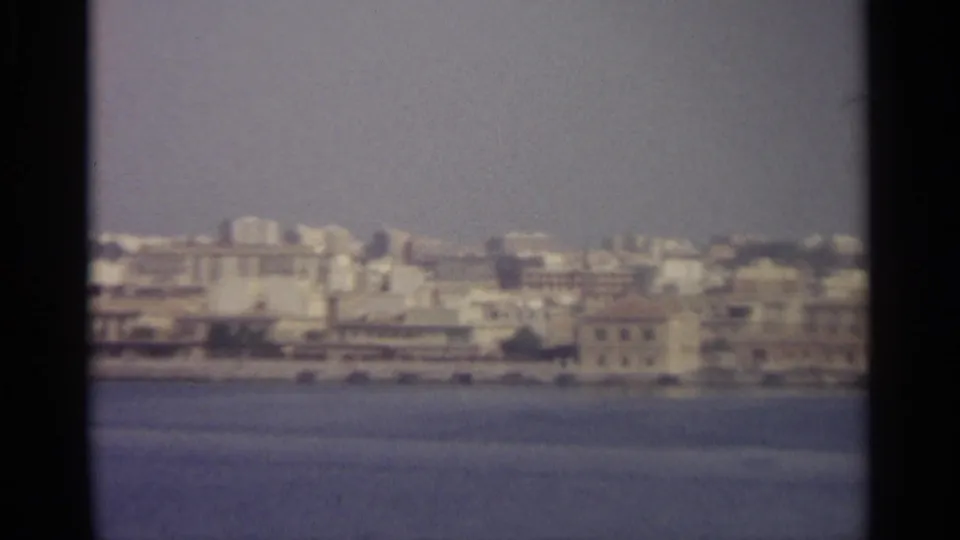Soft 8mm frame of a coastal Sicilian city from the water, with waterfront buildings and a distant tower visible.