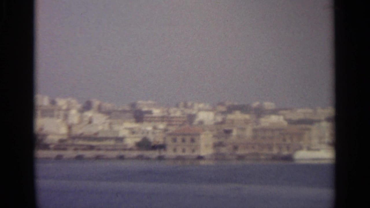 Soft 8mm view of a coastal town across the water, with dark frame borders and a hazy shoreline.