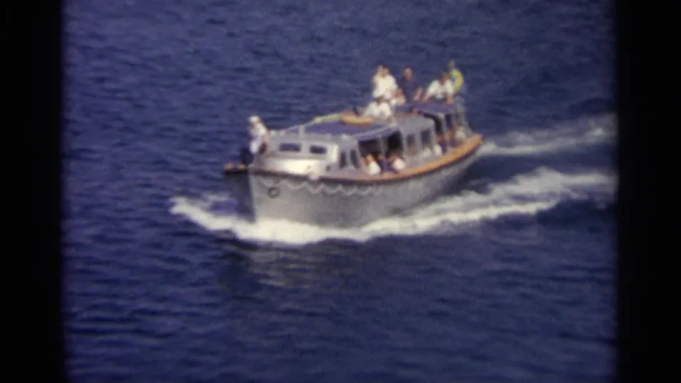 A small cabin motorboat with several visible passengers crosses deep blue water, leaving a white wake.
