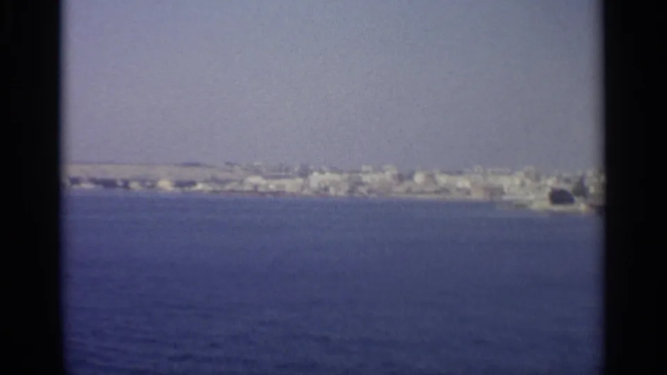 A distant pale shoreline town appears across blue water in a soft 8mm coastal view.