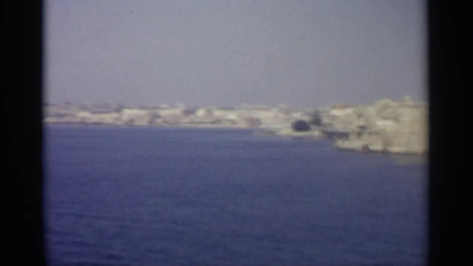 Soft 1965 8mm view across blue sea toward a pale Mediterranean coastal town, with a fort-like stone structure near the shoreline.
