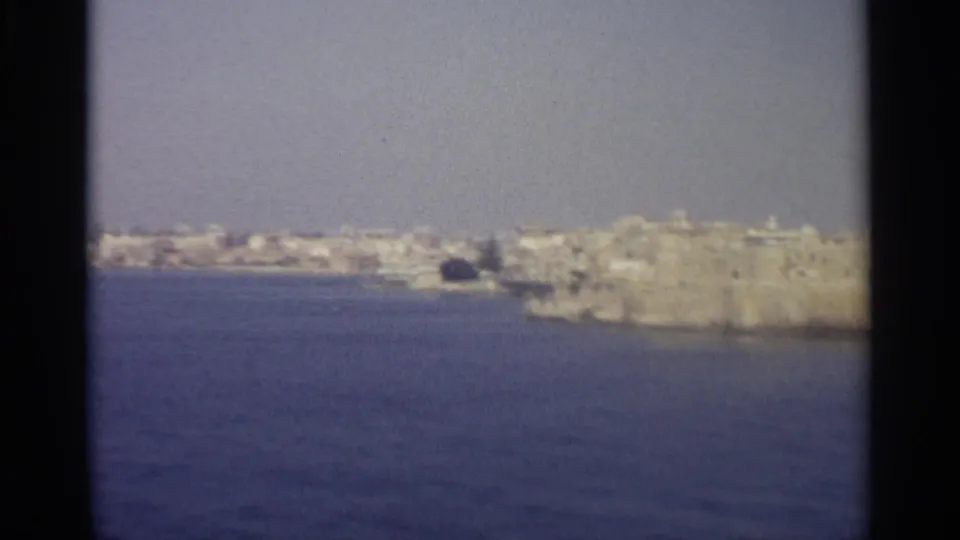 Distant 8mm frame of a pale coastal settlement and prominent stone waterfront mass viewed from across the sea.