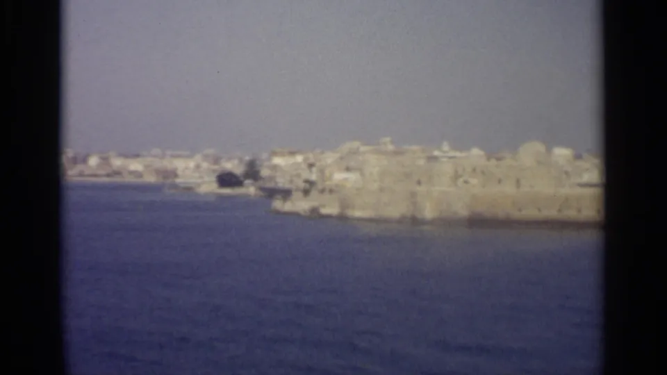 Vintage 8mm coastal view with deep blue water below a distant pale town and stone waterfront, softly framed by the film scan.