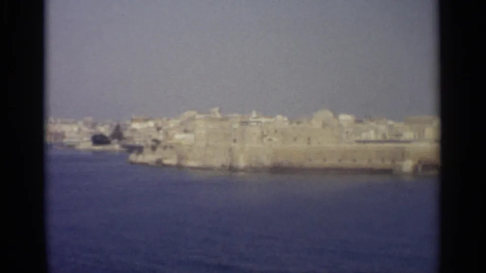 Soft 1965 home-movie frame of a Mediterranean-looking waterfront, with pale buildings and a broad stone fortification or wall above blue water.