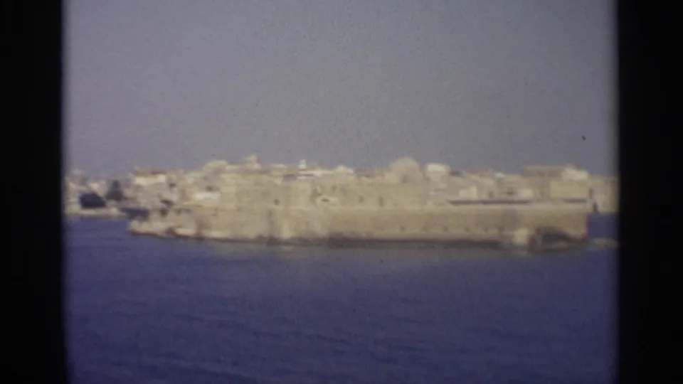 1965 8mm view of a historic coastal town, with stone waterfront walls and pale buildings seen across blue water.