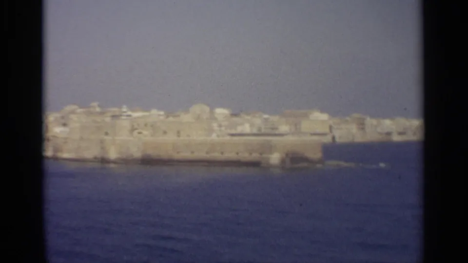 The camera holds on a fortified coastal edge with clustered pale buildings behind it, viewed from the water.