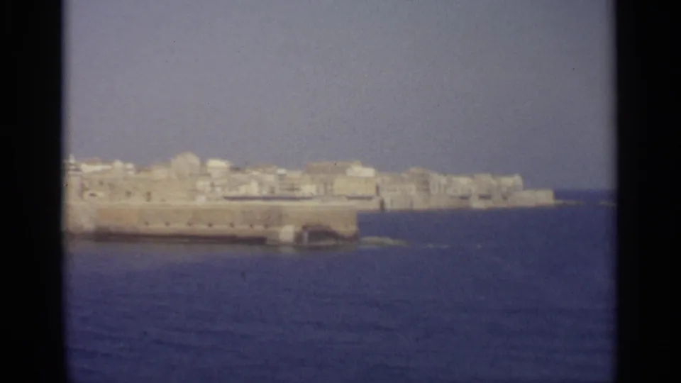 A drifting 8mm frame shows the stone waterfront and pale town buildings partly cropped as the shoreline continues rightward.