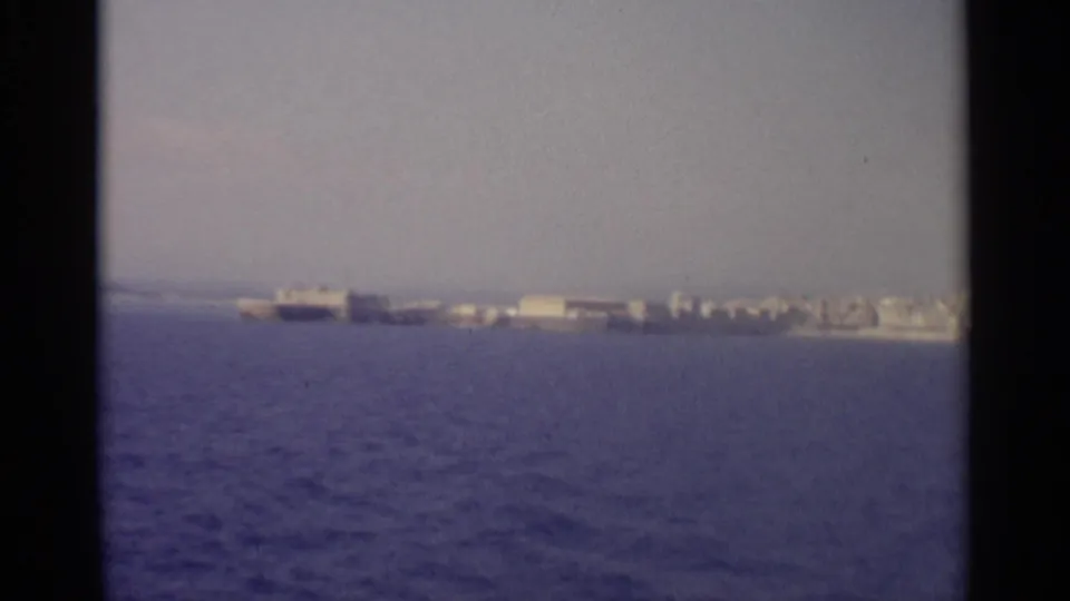 A distant coastal settlement appears across the water, with pale low-rise structures softened by 8mm focus and haze.