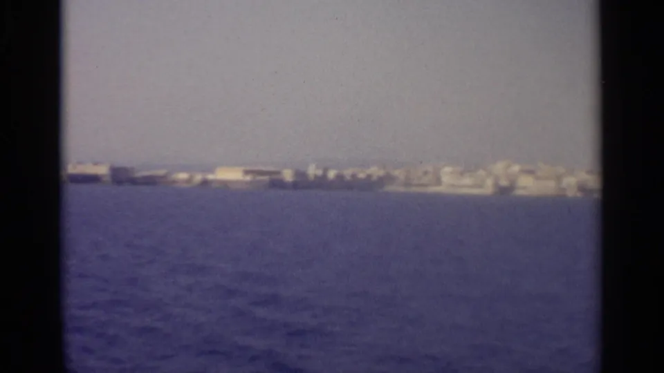 The boat draws nearer to a low coastal area with pale buildings along the shore, still distant and softly rendered.