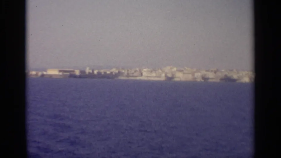 A wider coastal settlement is visible across blue water, framed by 8mm borders and haze from the moving-vessel viewpoint.