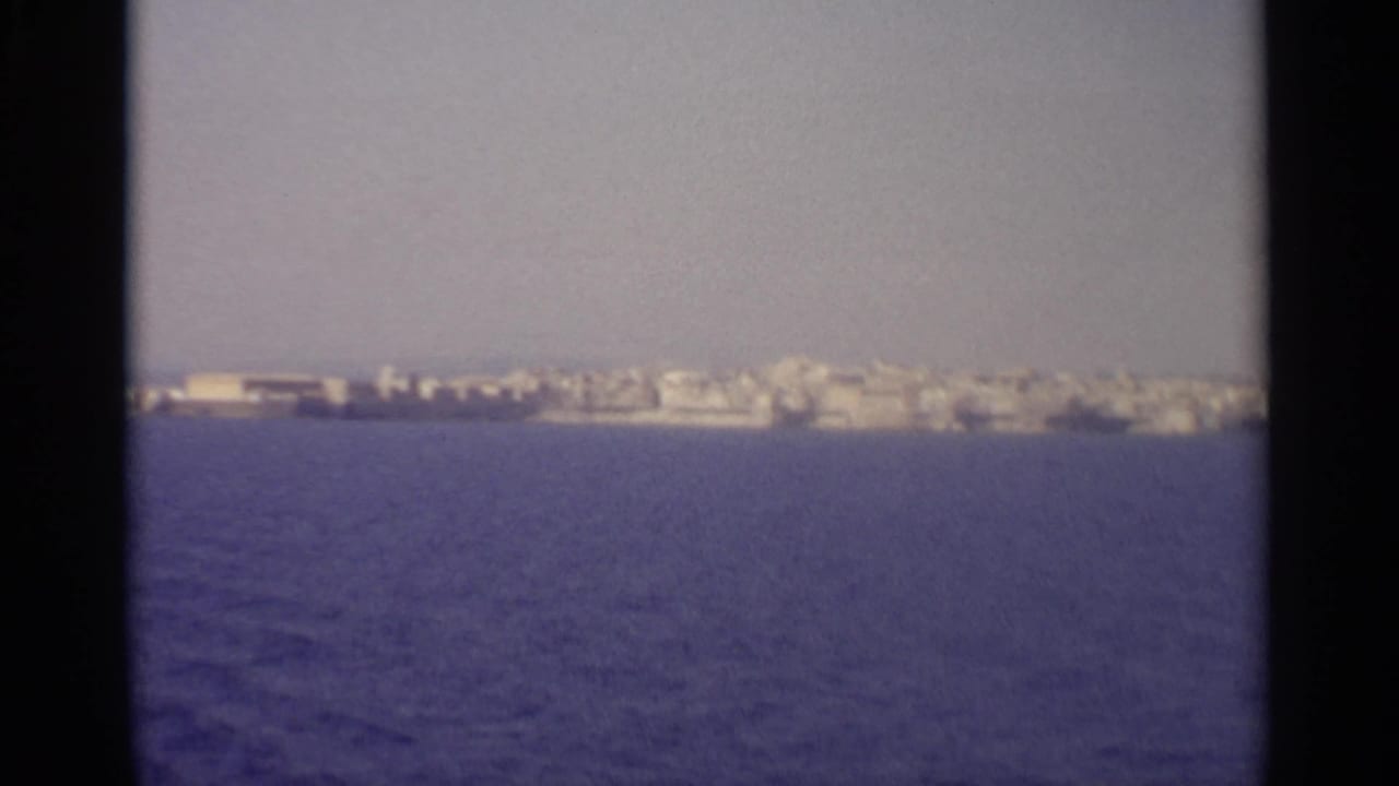 1965 8mm view from a boat toward a distant Sicilian shoreline with pale buildings and blue water.