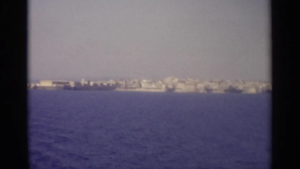 1965 8mm view from a boat toward a distant Sicilian shoreline with pale buildings and blue water.