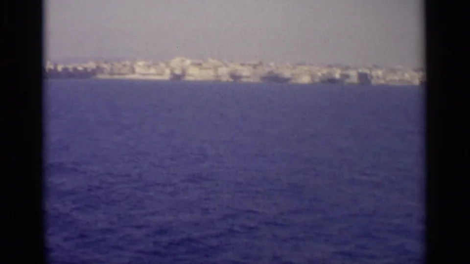 Distant Sicilian coastal buildings sit beyond deep blue water in a soft, grainy 8mm frame.