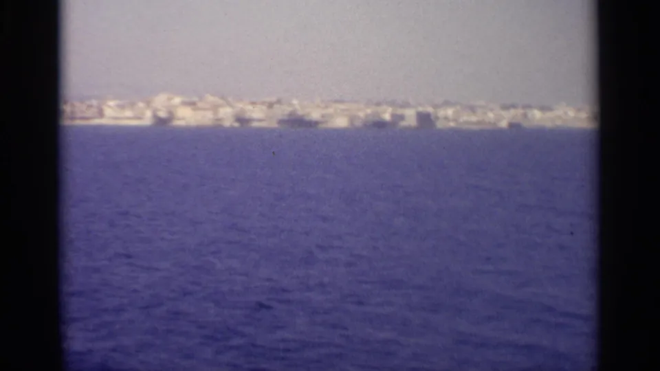 A 1965 home-movie frame shows the Sicilian shoreline stretching across the horizon over rippled sea.