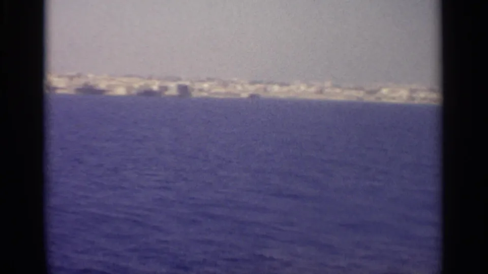 Distant coastal strip appears across open water in a soft 8mm frame, with pale buildings and shoreline forms under a hazy sky.