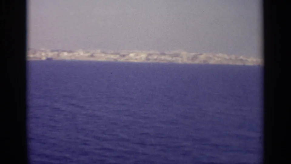 The frame shows a long distant shoreline with pale buildings above a wide band of blue sea, filmed in a handheld 8mm travel style.