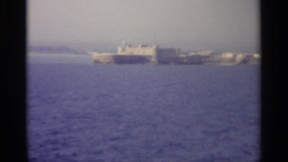 A large ship appears offshore near a distant hazy coastline, with blue sea filling the foreground in this 1965 8mm travel frame.