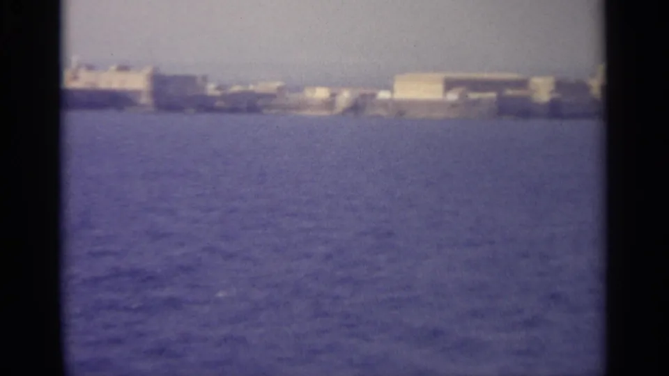 Grainy 8mm view of pale waterfront structures and low buildings on a distant coastal edge.