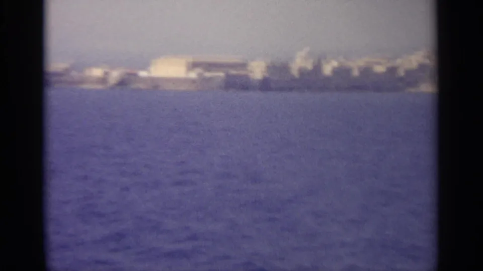 Vintage 8mm frame with blue water in the foreground and an indistinct coastal settlement beyond.