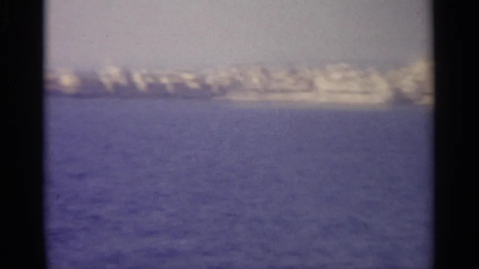 Soft, grainy 8mm view of clustered waterfront buildings or boats along a distant coastal shore.