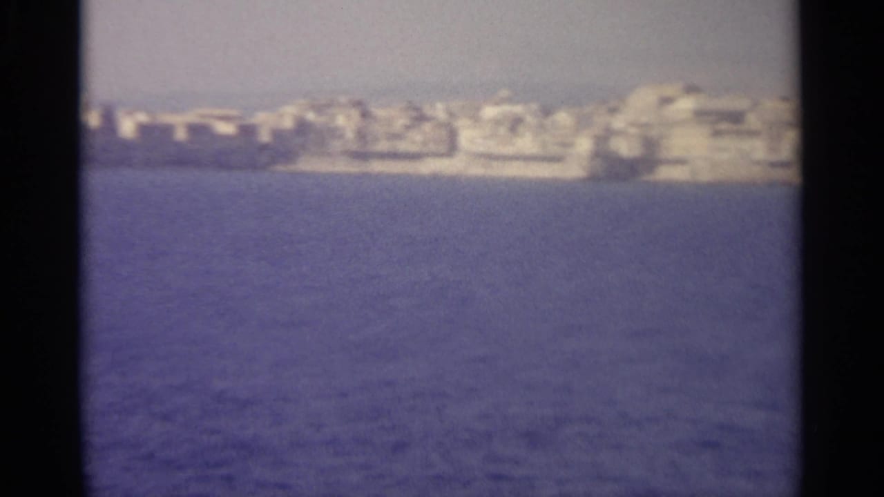 1965 8mm frame showing a soft distant Sicilian coastal settlement across deep blue sea with black film borders.