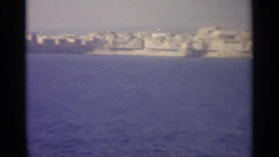 1965 8mm frame showing a soft distant Sicilian coastal settlement across deep blue sea with black film borders.
