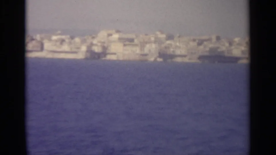 Soft 8mm view of pale Sicilian coastal buildings on the horizon beyond blue water and dark frame edges.