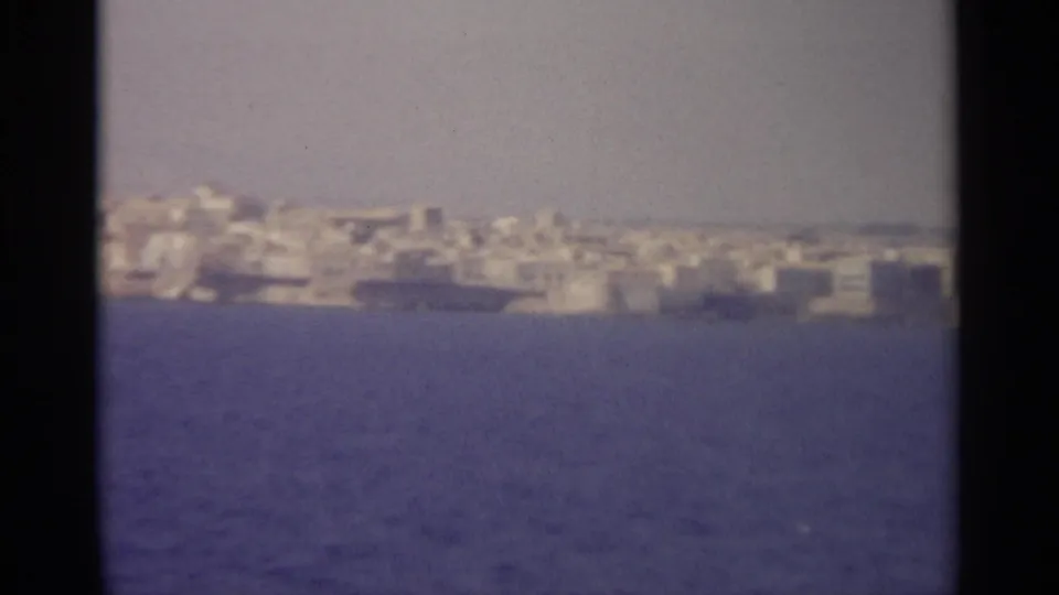 1965 home-movie frame with a distant Sicilian waterfront settlement, soft focus, grain, and deep blue water foreground.