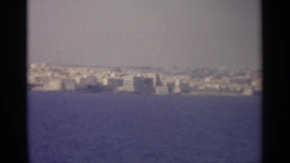 Aged 8mm frame showing a distant Sicilian coastal town below a low hill, viewed from across the sea.