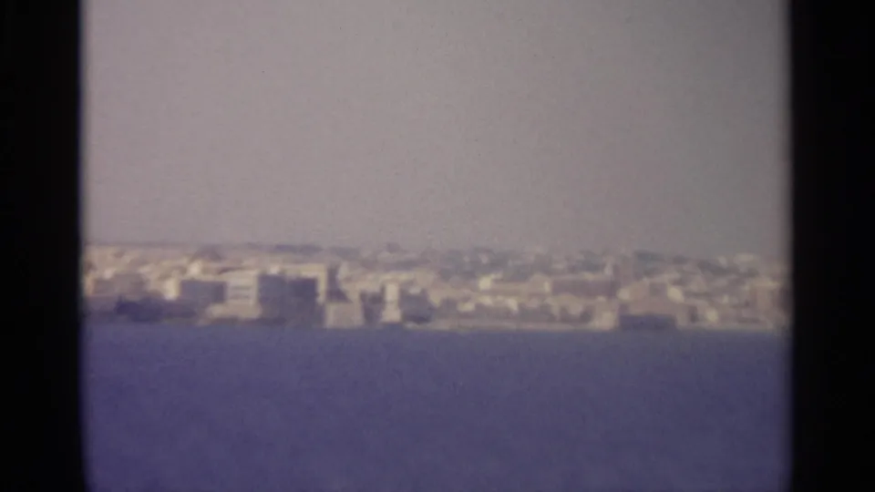 Soft 8mm frame of a distant coastal cityscape across calm water, described as Sicily in 1965 by clip metadata.