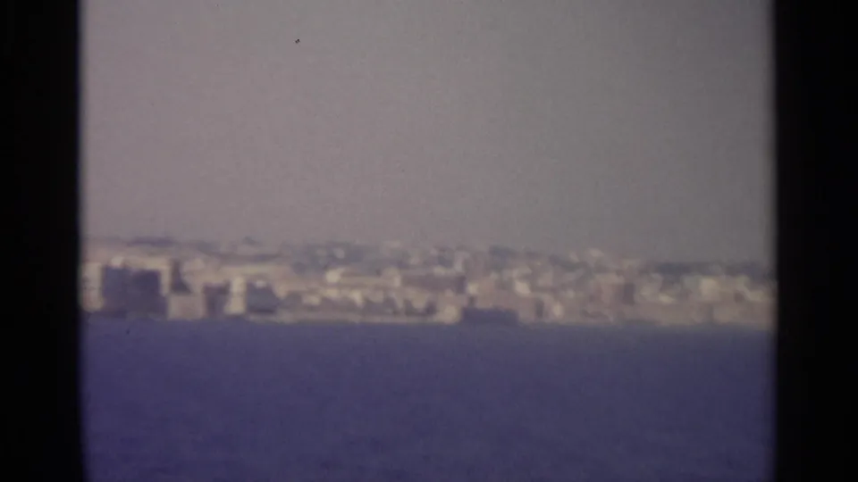 Distant low-rise coastal buildings sit beyond blue water in a hazy 8mm travel view, with a dark gate edge visible.