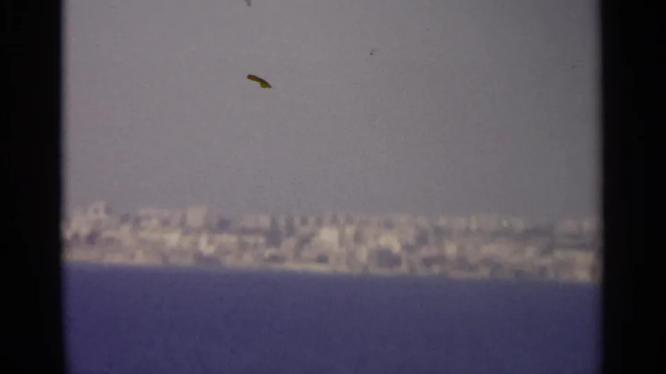 A grainy 8mm view looks across the sea toward a pale, distant coastal cityscape with no visible people or signage.