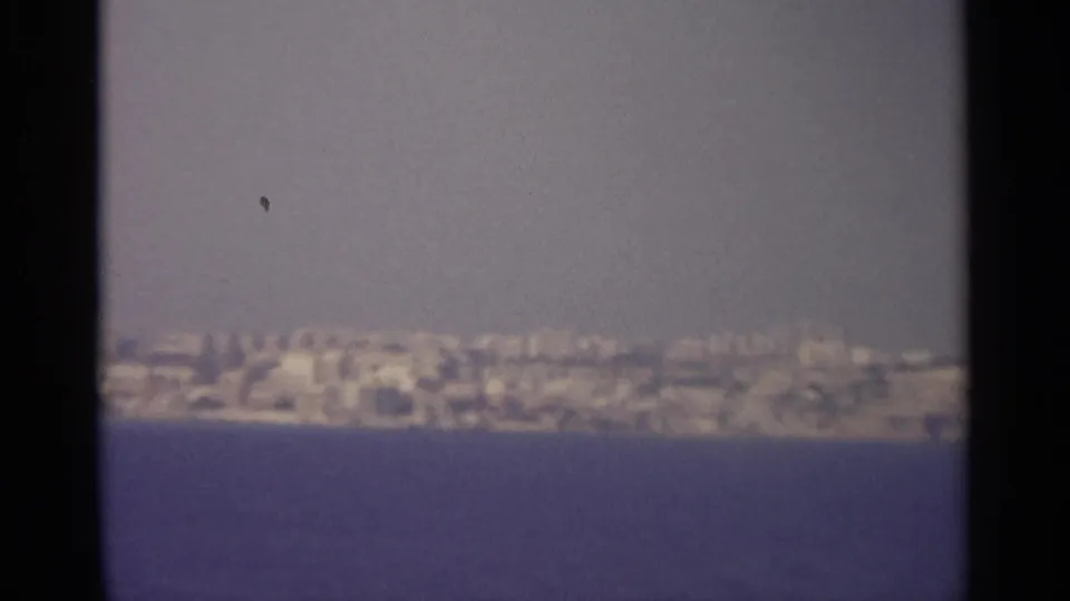 1960s 8mm home-movie view of a distant coastal settlement across blue water, seen through soft focus and film grain.