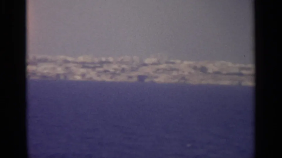 Distant coastal buildings and land sit beyond open blue water in faded 8mm color, with the frame edge visible at the side.