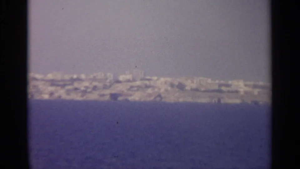 Soft 8mm frame of a hazy coastal horizon, with pale buildings along the shore and deep blue water in the foreground.