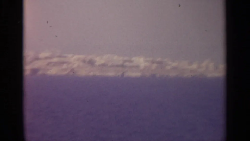 Faded-color 8mm view of a distant shoreline under a pale pink sky, with water filling the lower half of the frame.