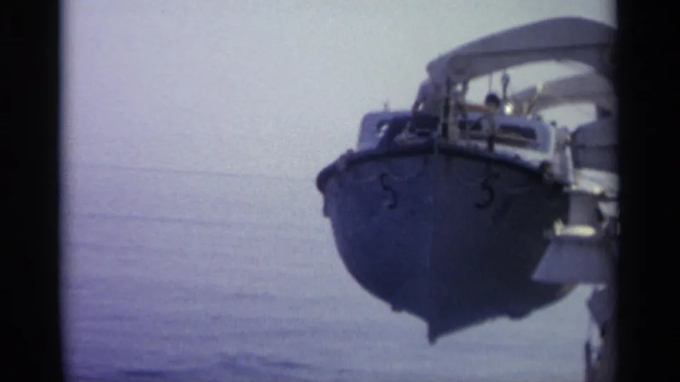 Small military-looking boat with bow number 5, railings, and canopy structure seen on calm water under a hazy sky.