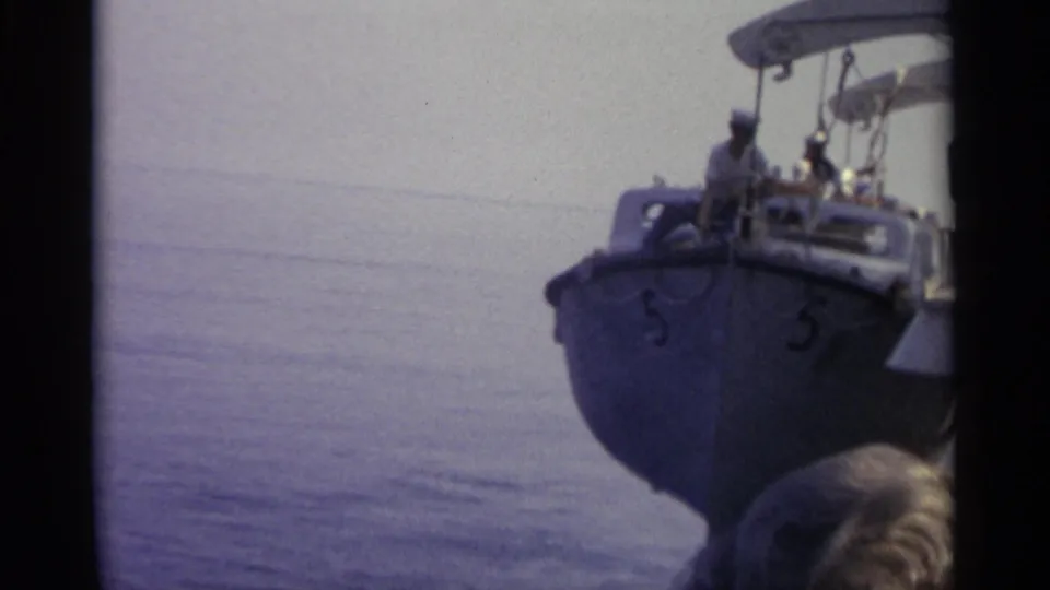 A small blue boat carrying passengers appears beside a nearby vessel, with a partial head in the foreground and calm water beyond.