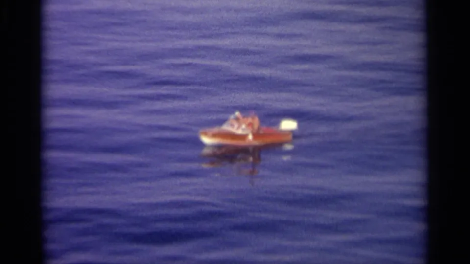 A distant motorboat with visible occupants floats on rippled blue water, framed by soft 8mm film texture.