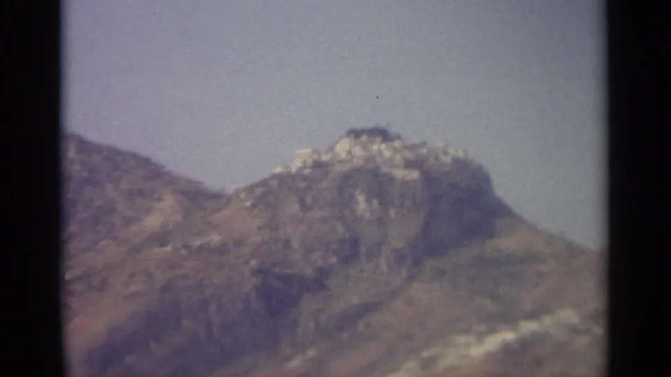 A rugged mountain slope with distant pale buildings appears under a hazy sky in the 1965 home-movie sequence.