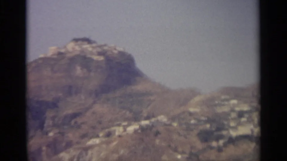 A closer view of the rugged hillside shows scattered light-colored buildings against a muted, hazy sky.