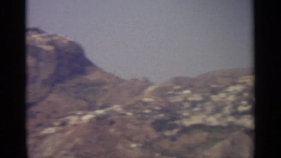 1960s 8mm frame of a dry mountain slope with scattered pale buildings and soft, faded color.