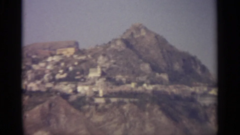 Faded 1960s home-movie frame showing a rugged peak above a distant hillside settlement.